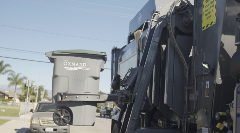 An Oxnard trash truck picking up a trash can with the City of Oxnard logo on it.